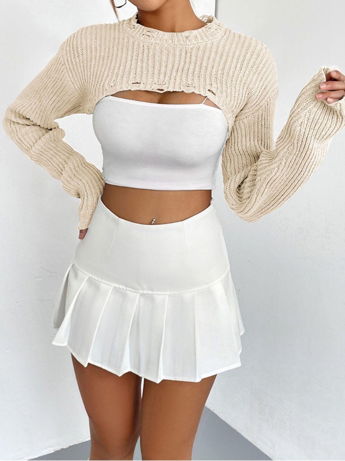woman wearing a white crop top, a beige cropped sweater with cut-out details, and a white pleated mini skirt