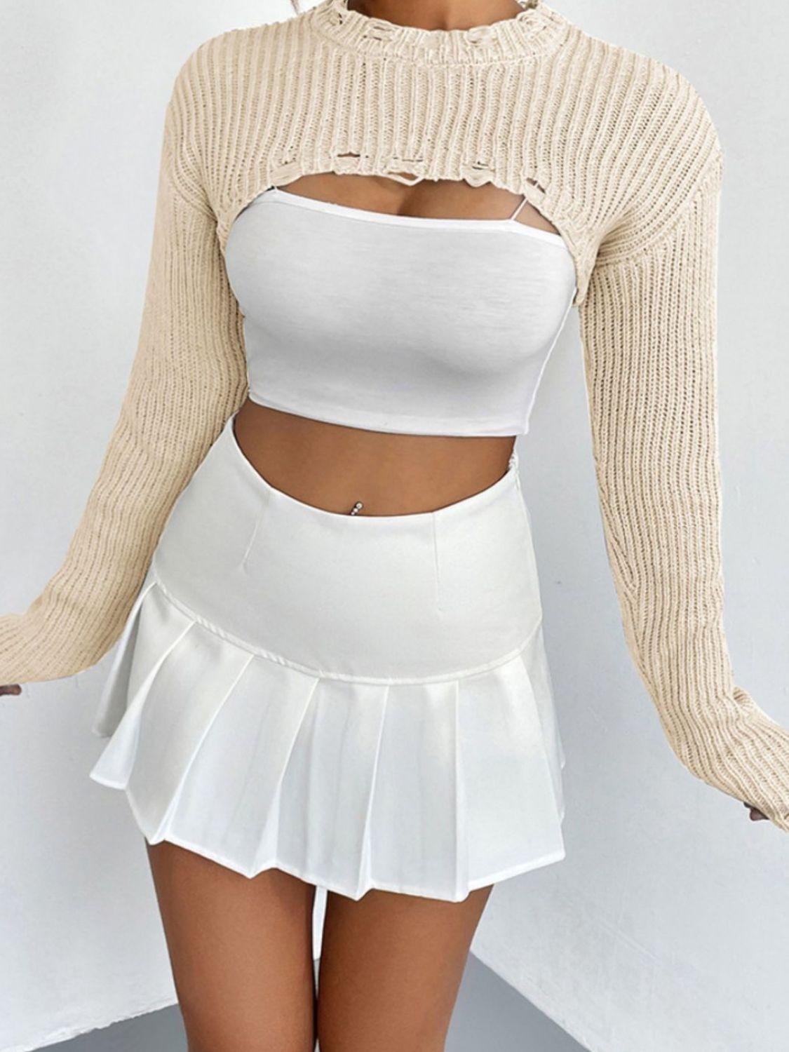 woman wearing a white crop top and a beige cropped sweater with distressed details, and a white pleated mini skirt