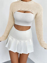 woman wearing a white crop top and a beige cropped sweater with distressed details, and a white pleated mini skirt