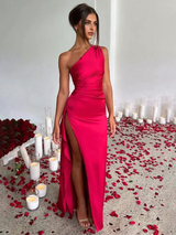 Woman in a fuchsia one-shoulder satin gown with a high slit, standing in a room decorated with candles and rose petals.