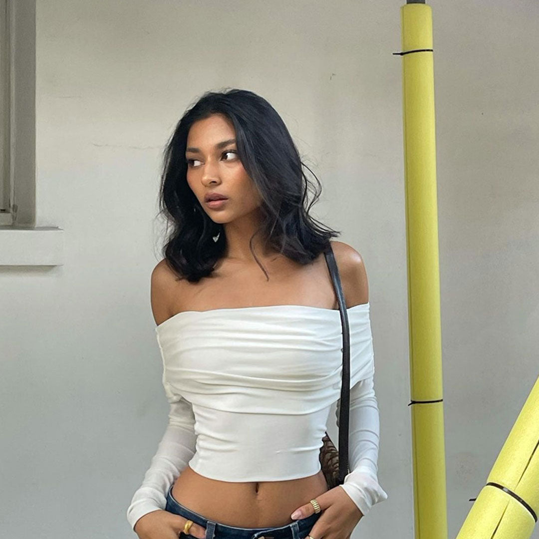 A woman wearing a white off-the-shoulder top and blue jeans stands next to a yellow pole.