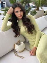 A young woman with long dark wavy hair and blue eyes poses in a lime green ribbed knit maxi dress, leaning on a white sofa.