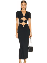 A woman stands facing forward wearing a black, floor-length dress with short sleeves and cutouts at the waist and chest, accented with gold rings. She holds a small black handbag and wears gold earrings.