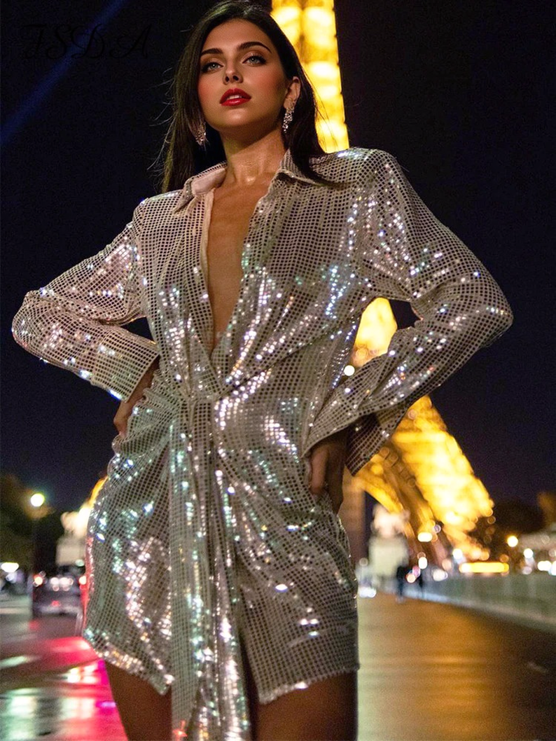 Woman wearing a sparkling silver sequined dress with a plunging neckline and tie waist, posing in front of the Eiffel Tower at night.