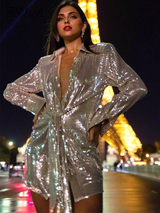 Woman wearing a sparkling silver sequined dress with a plunging neckline and tie waist, posing in front of the Eiffel Tower at night.