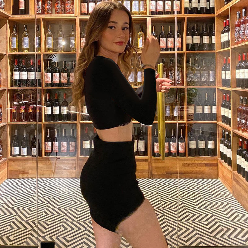 Young woman in a black crop top and mini skirt with silver fringe detail, posing in front of a wall of wine bottles.