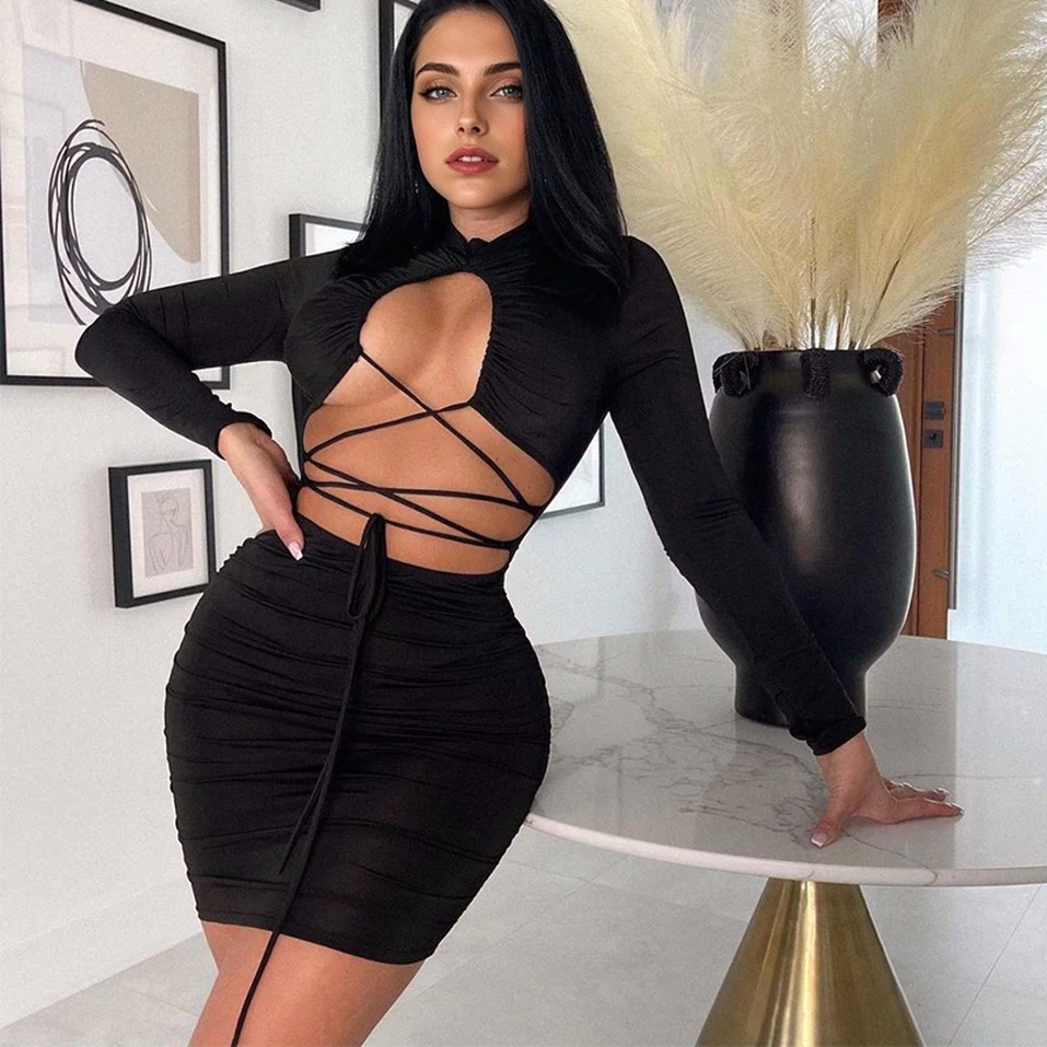 Woman in a black long-sleeved, cut-out, lace-up, ruched mini dress leaning on a table.