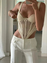 A woman in a cream knit top with a crochet overlay and white pants, holding her hand to her chest.