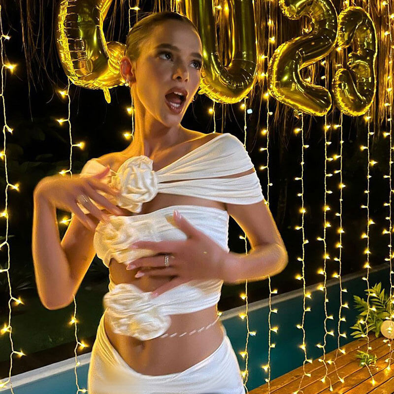A woman in a white off-the-shoulder top with floral embellishments and matching high-waisted pants poses with her hands near her chest.