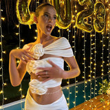 A woman in a white off-the-shoulder top with floral embellishments and matching high-waisted pants poses with her hands near her chest.
