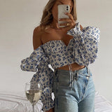 Woman taking a mirror selfie, wearing a white and blue floral print off-the-shoulder cropped top with dramatic puff sleeves and light blue criss-cross high-waisted jeans, with a glass of white wine on the bed.