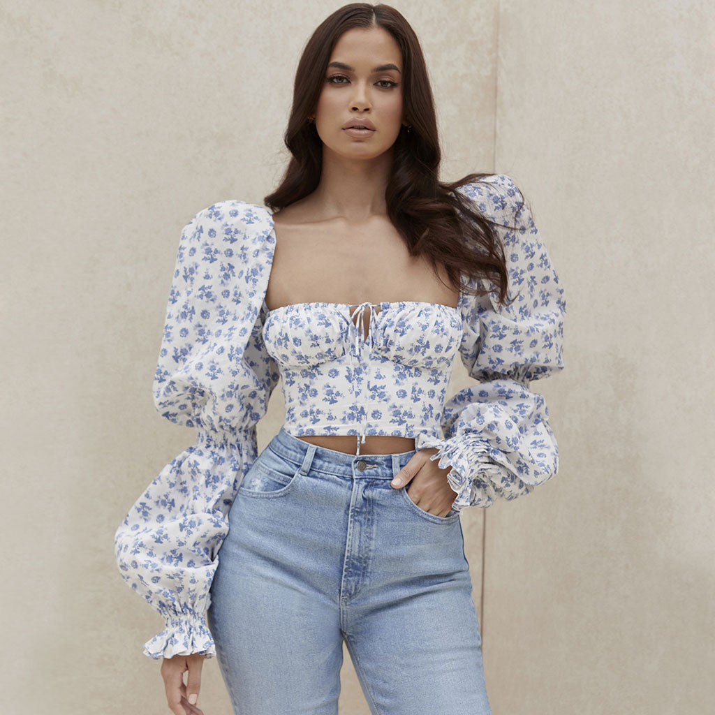 Woman wearing a white and blue floral print cropped top with puff sleeves and light blue high-waisted jeans, standing against a beige wall.