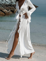 A woman in a white sheer maxi dress with ruffled trim and a high slit walks on a sandy beach with clear blue water.