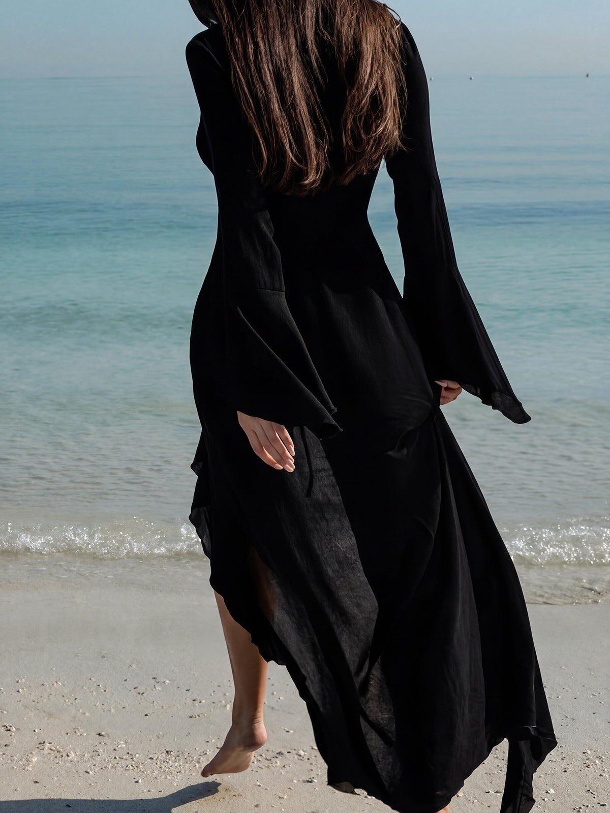 A woman from behind in a black sheer maxi dress with flared sleeves walks on the beach, the fabric flowing behind her.