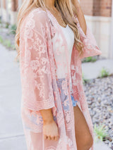 A woman wearing a pink lace kimono over a white tank top and denim shorts.