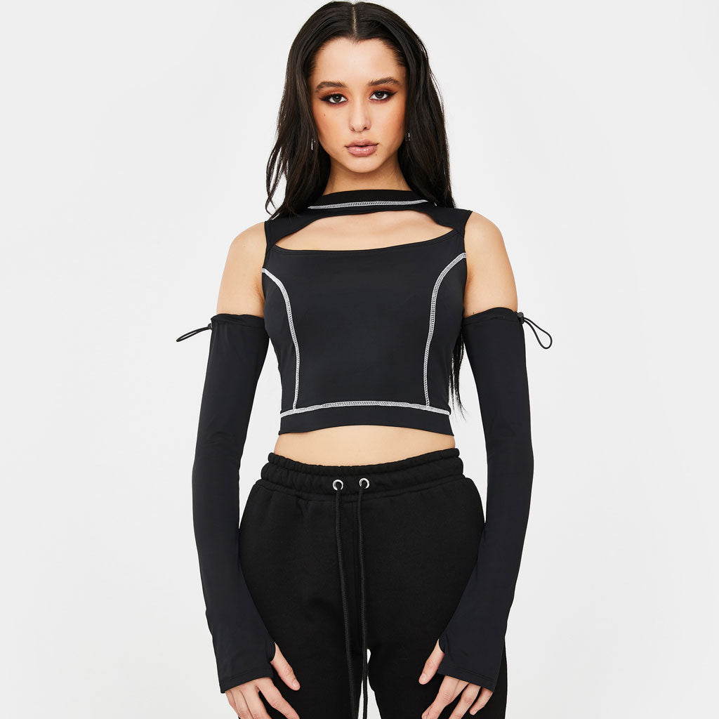 A young woman with long dark hair stands against a white background, wearing a black cropped top with a cut-out neckline and white contrast stitching. She also wears long black sleeves that extend from the shoulders and black sweatpants.