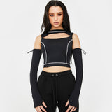A young woman with long dark hair stands against a white background, wearing a black cropped top with a cut-out neckline and white contrast stitching. She also wears long black sleeves that extend from the shoulders and black sweatpants.