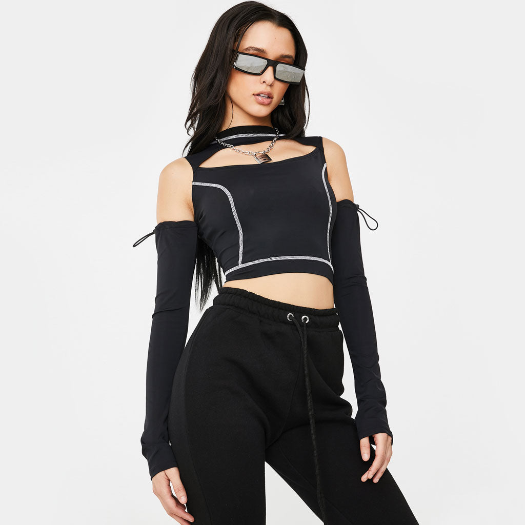 A young woman with long dark hair and sunglasses poses against a white background. She is wearing a black cropped top with a cut-out neckline and white contrast stitching, long black sleeves, and black sweatpants.