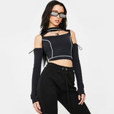 A young woman with long dark hair and sunglasses poses against a white background. She is wearing a black cropped top with a cut-out neckline and white contrast stitching, long black sleeves, and black sweatpants.