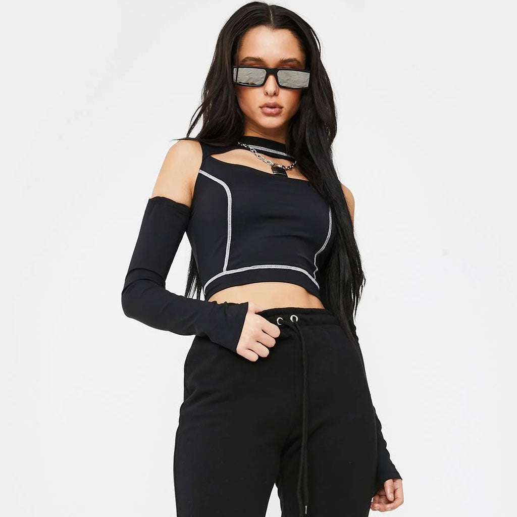 A young woman with long dark hair and sunglasses poses against a white background. She is wearing a black cropped top with a cut-out neckline and white contrast stitching, long black sleeves, and black sweatpants. She is looking to the side.