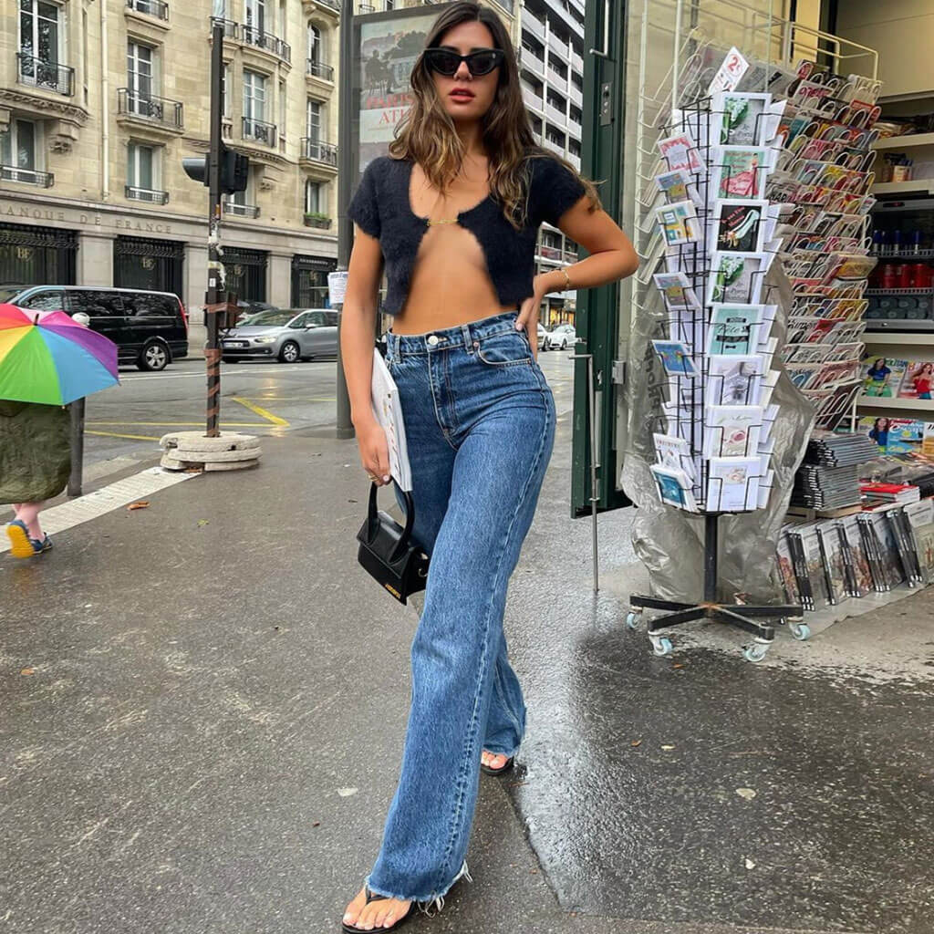 A woman in a black fuzzy cropped cardigan and blue jeans stands on a city sidewalk.