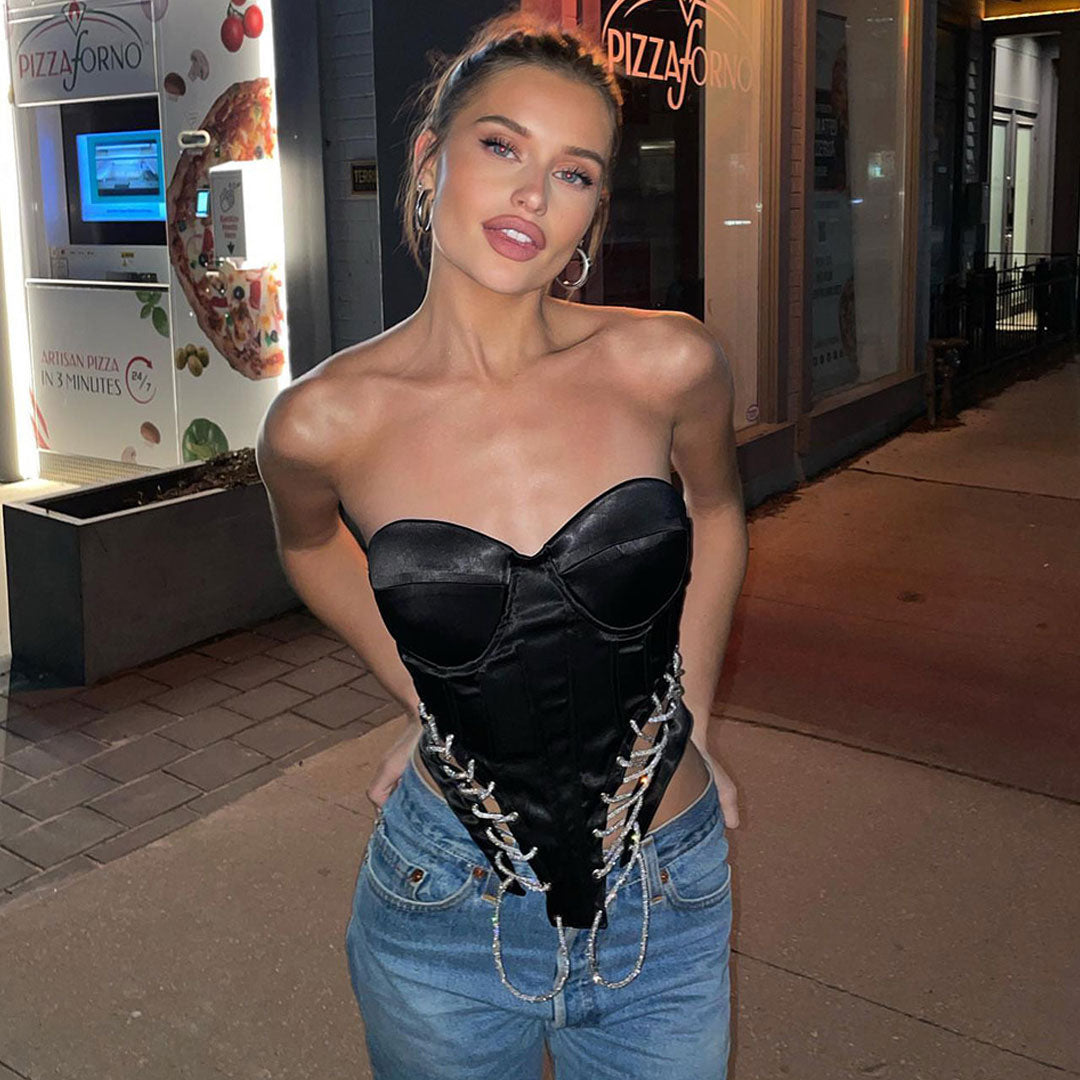 A woman wearing a black strapless corset top with lace-up and chain detailing, paired with blue jeans, standing on a sidewalk at night.