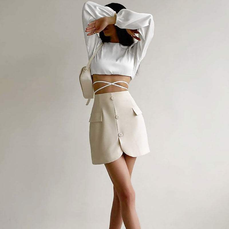 woman wearing a white cropped top and a beige skirt with her arms covering her face