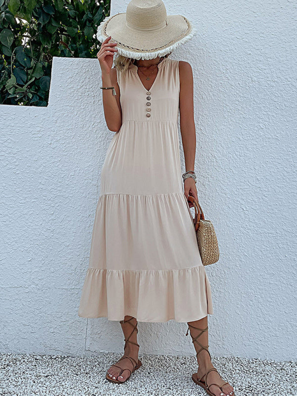 woman in a beige tiered midi dress, straw hat, and straw bag, standing against a white wall