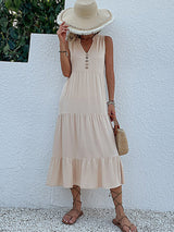 woman in a beige tiered midi dress, straw hat, and straw bag, standing against a white wall
