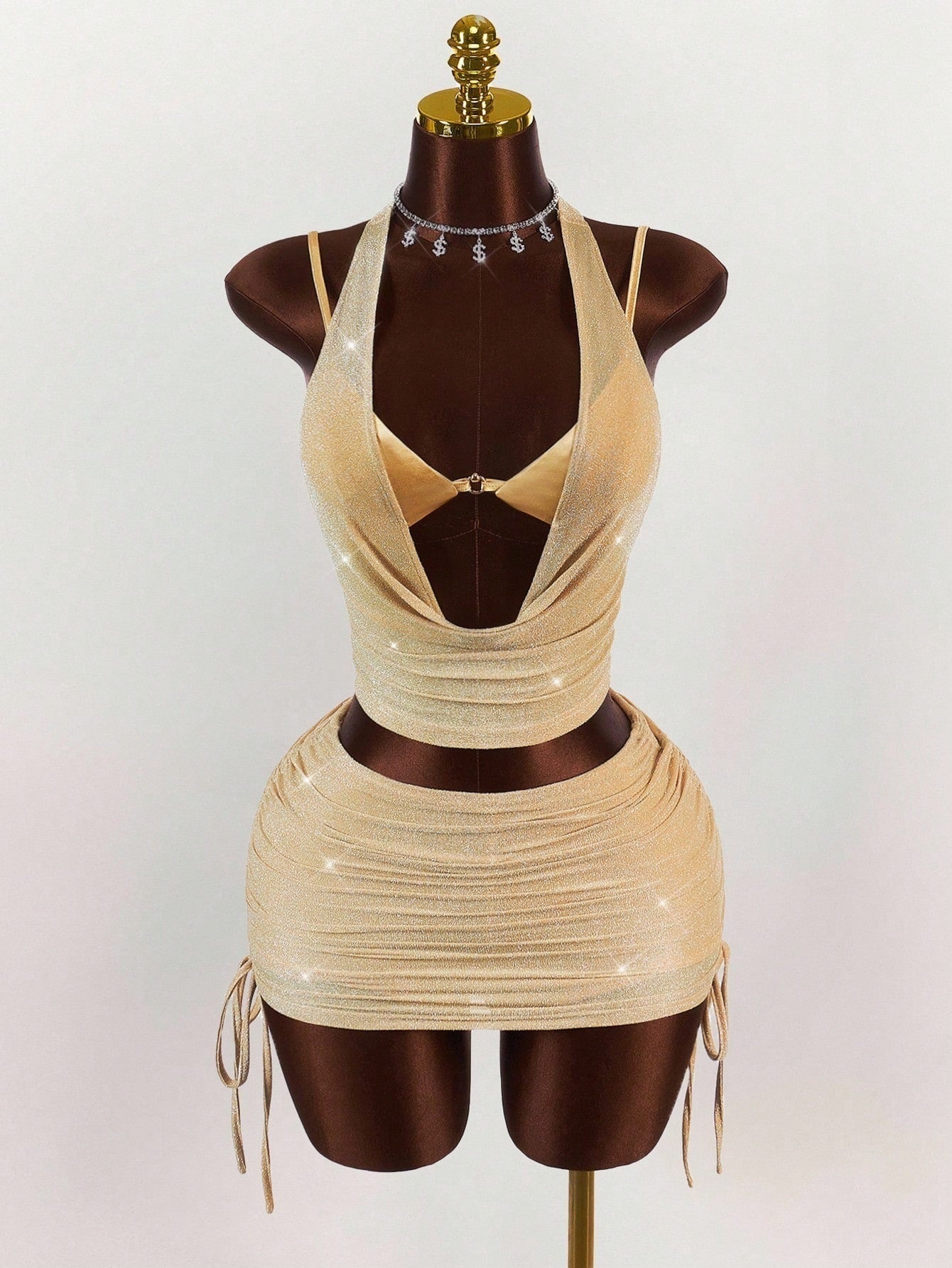 A gold sparkly halter neck crop top and ruched mini skirt set on a mannequin, accessorized with a dollar sign necklace.