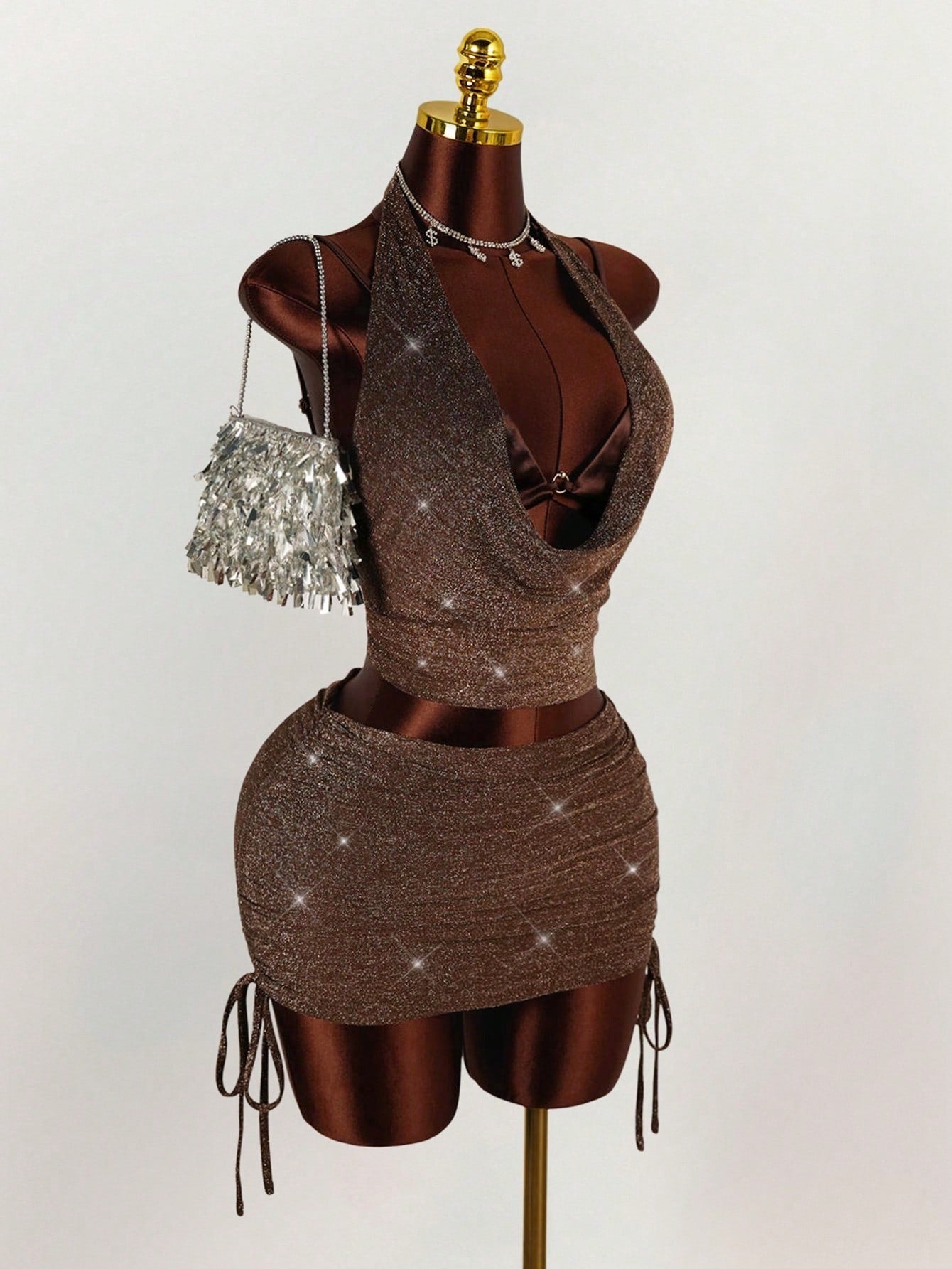 A brown sparkly halter neck crop top and ruched mini skirt set with a drawstring detail on the skirt, shown with a silver fringe purse and dollar sign necklace.
