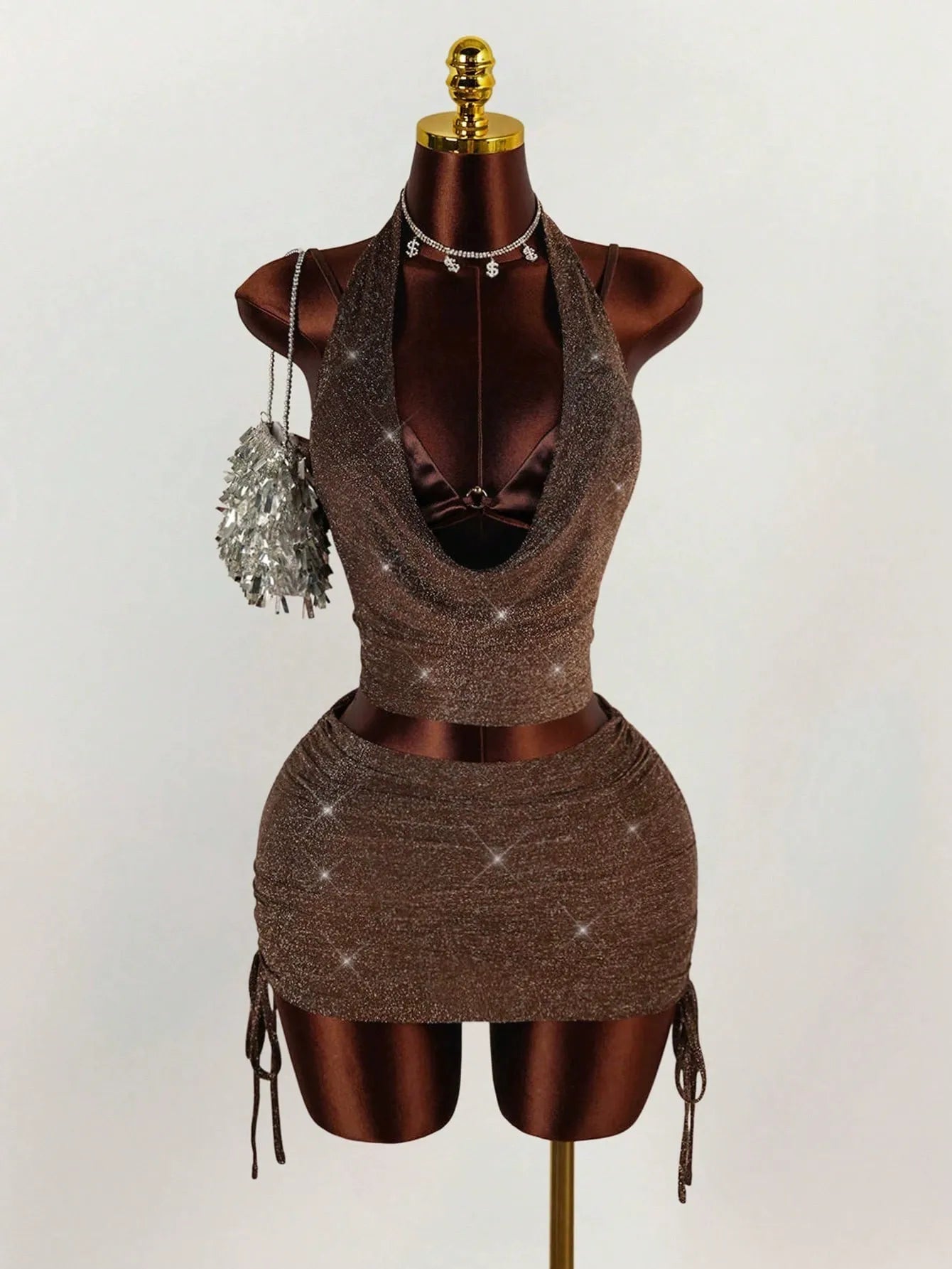 A brown sparkly halter neck crop top and ruched mini skirt set on a mannequin, accessorized with a silver fringe purse and a dollar sign necklace.