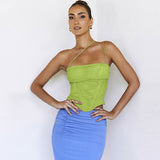 A woman stands with her hands on her hips against a white wall, wearing a lime green asymmetrical strap top and a blue ruched skirt.