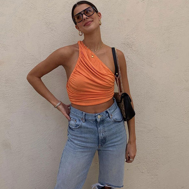 Woman wearing an orange one-shoulder ruched crop top and light wash ripped jeans, posing against a beige wall.