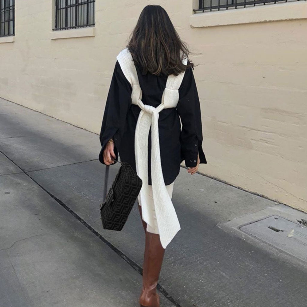 A woman from behind wearing a white knit wrap top tied over a black shirt, with white pants and brown boots.