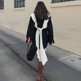 A woman from behind wearing a white knit wrap top tied over a black shirt, with white pants and brown boots.