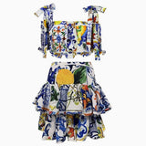 A white two-piece set with a colorful blue, yellow, green, and orange tile and floral print. The top is cropped with a square neckline and tie shoulder straps. The skirt is a mini skirt with ruffles.