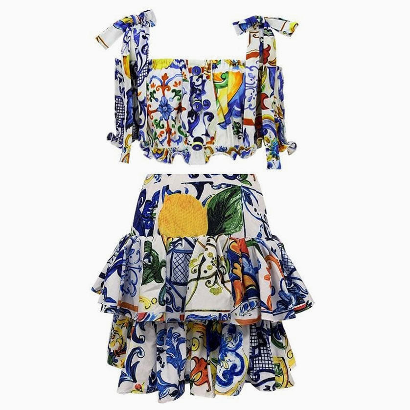 A white two-piece set with a colorful blue, yellow, green, and orange tile and floral print. The top is cropped with a square neckline and tie shoulder straps. The skirt is a mini skirt with ruffles.