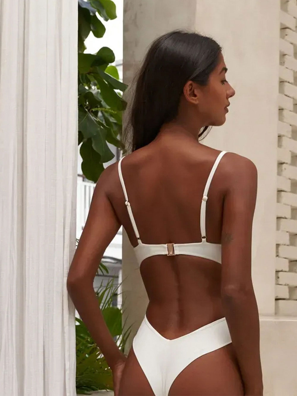 A woman wearing a cream one-piece swimsuit, viewed from the back, showing the scoop back and adjustable straps.