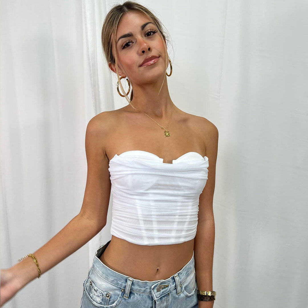 A young woman in a white strapless ruched top and light blue jeans poses in front of a white curtain.