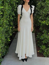 A woman stands on a pathway surrounded by lush green bushes, wearing a long white dress with black ribbon accents on the shoulders.