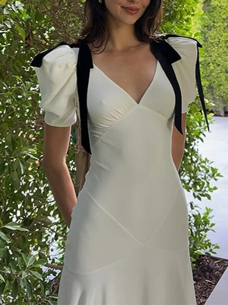 A woman's upper body is shown in a white dress with puff sleeves and black ribbon details on the shoulders, with a blurred green background.