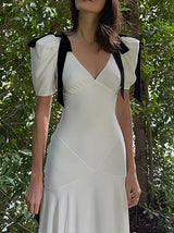 A close-up of a woman wearing a white dress with puff sleeves and black ribbon details on the shoulders, set against a backdrop of green trees.
