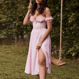 Woman wearing a pink floral off-the-shoulder midi dress with a high slit, standing next to a swing set outdoors.