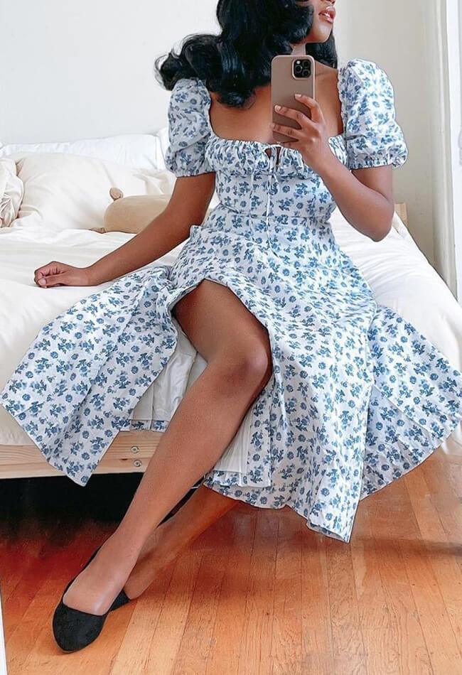 Woman with dark curly hair sitting on a bed wearing a white and blue floral print dress with a square neckline, puff sleeves, and a high slit.