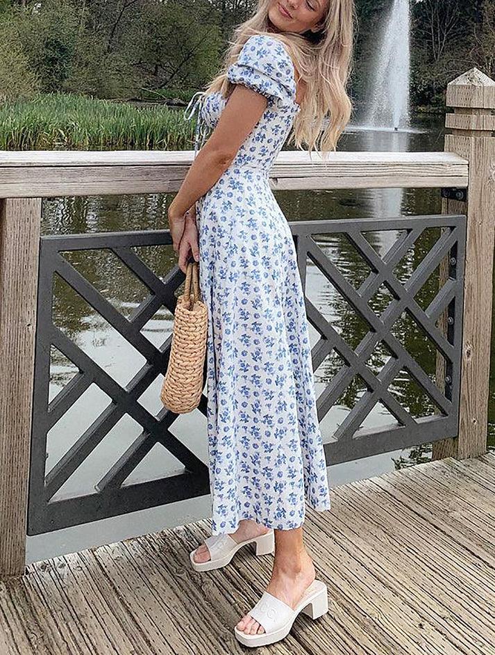 Woman in a white and blue floral print dress with a square neckline, puff sleeves, and a high slit, standing on a wooden bridge.