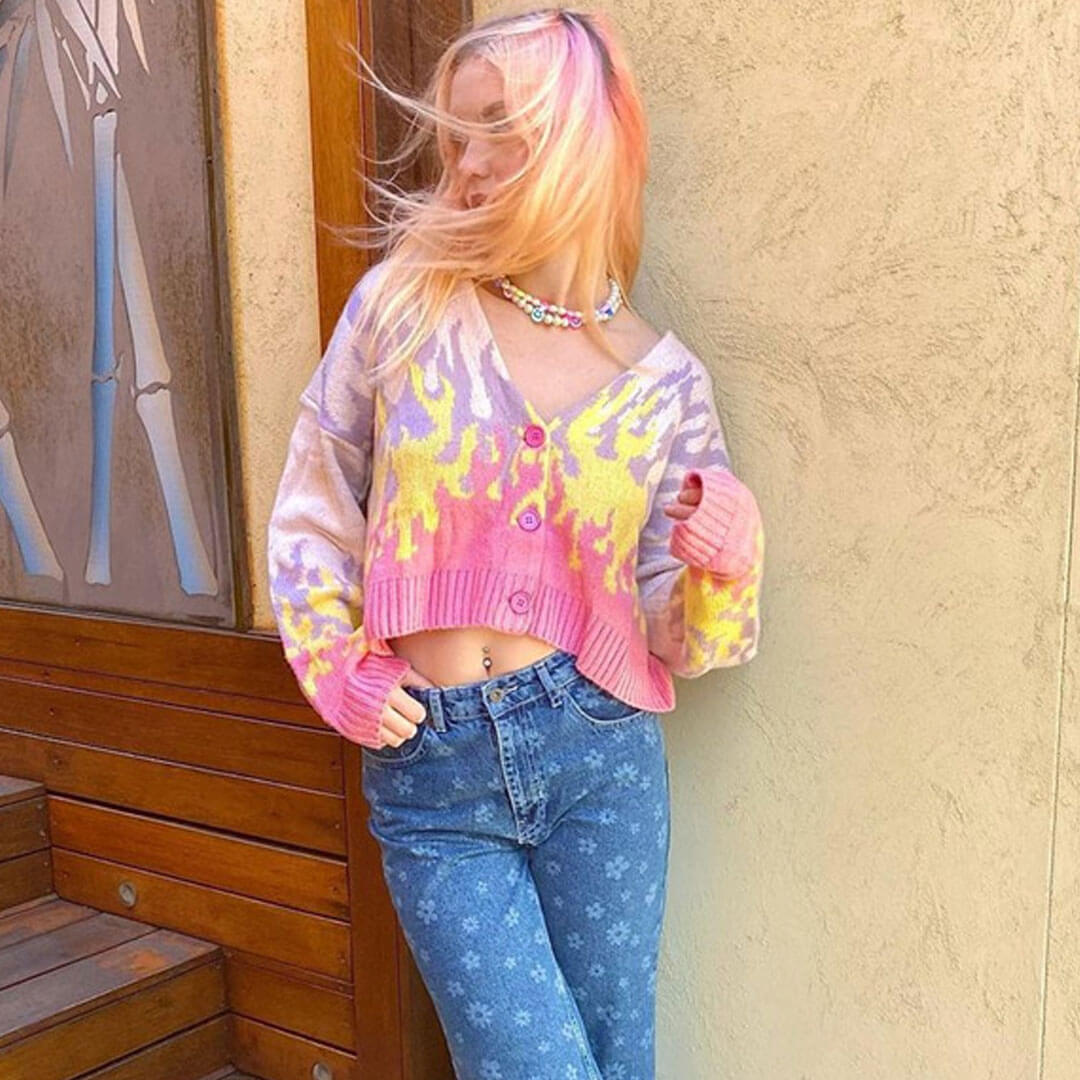 A young woman with pink hair looking to the side, her hair blowing in the wind, wearing a cropped flame print cardigan and blue jeans.
