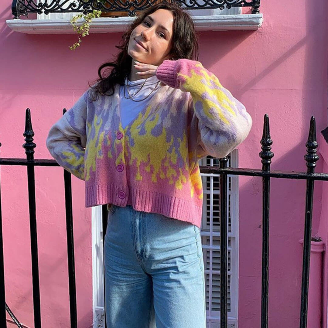 A young woman with dark hair wearing a cropped flame print cardigan over a white top and light blue jeans, posing in front of a pink building.