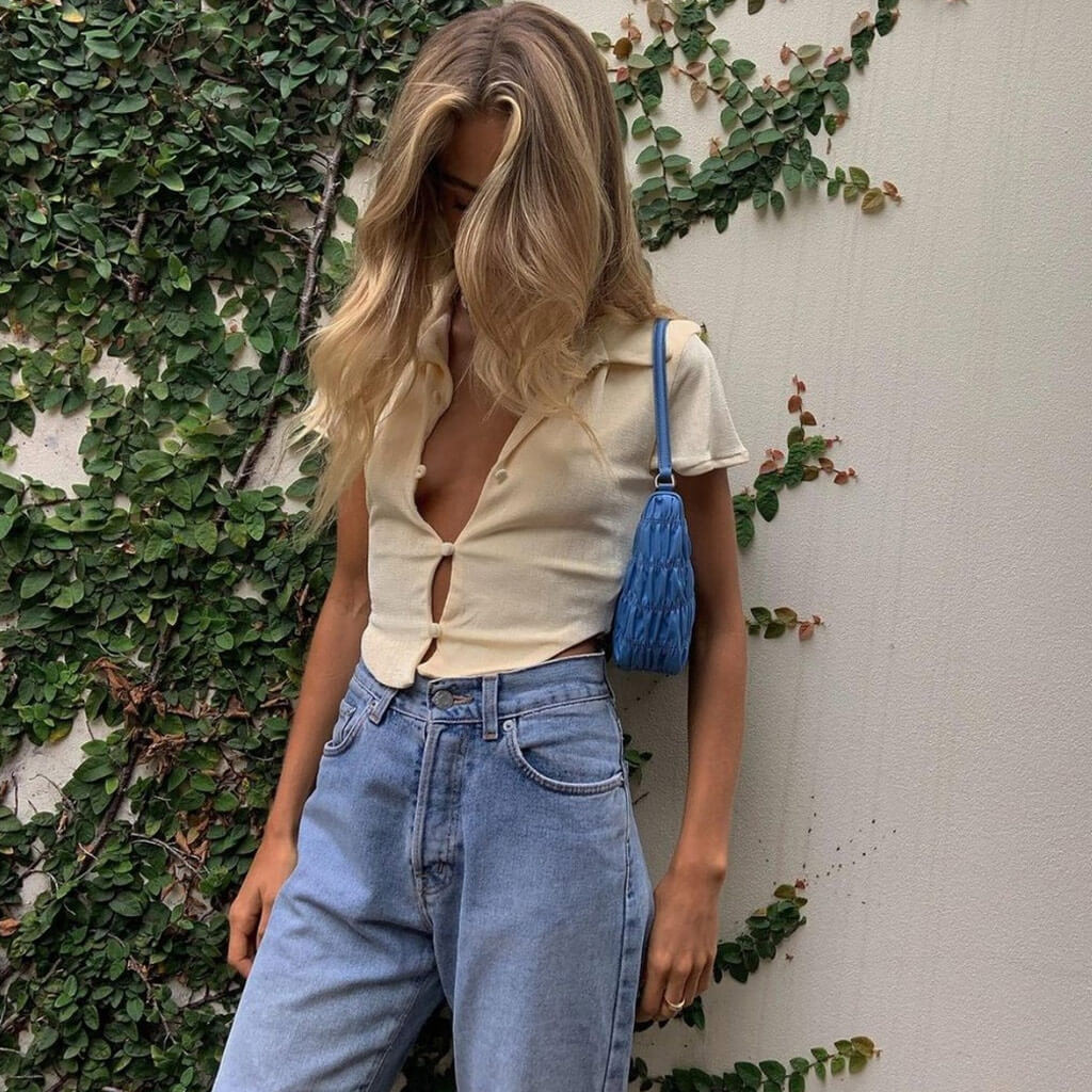 A woman with blonde hair down her back stands against a wall covered in ivy, wearing a cream-colored cropped collared shirt, blue jeans, and carrying a blue purse.