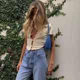A woman with blonde hair down her back stands against a wall covered in ivy, wearing a cream-colored cropped collared shirt, blue jeans, and carrying a blue purse.